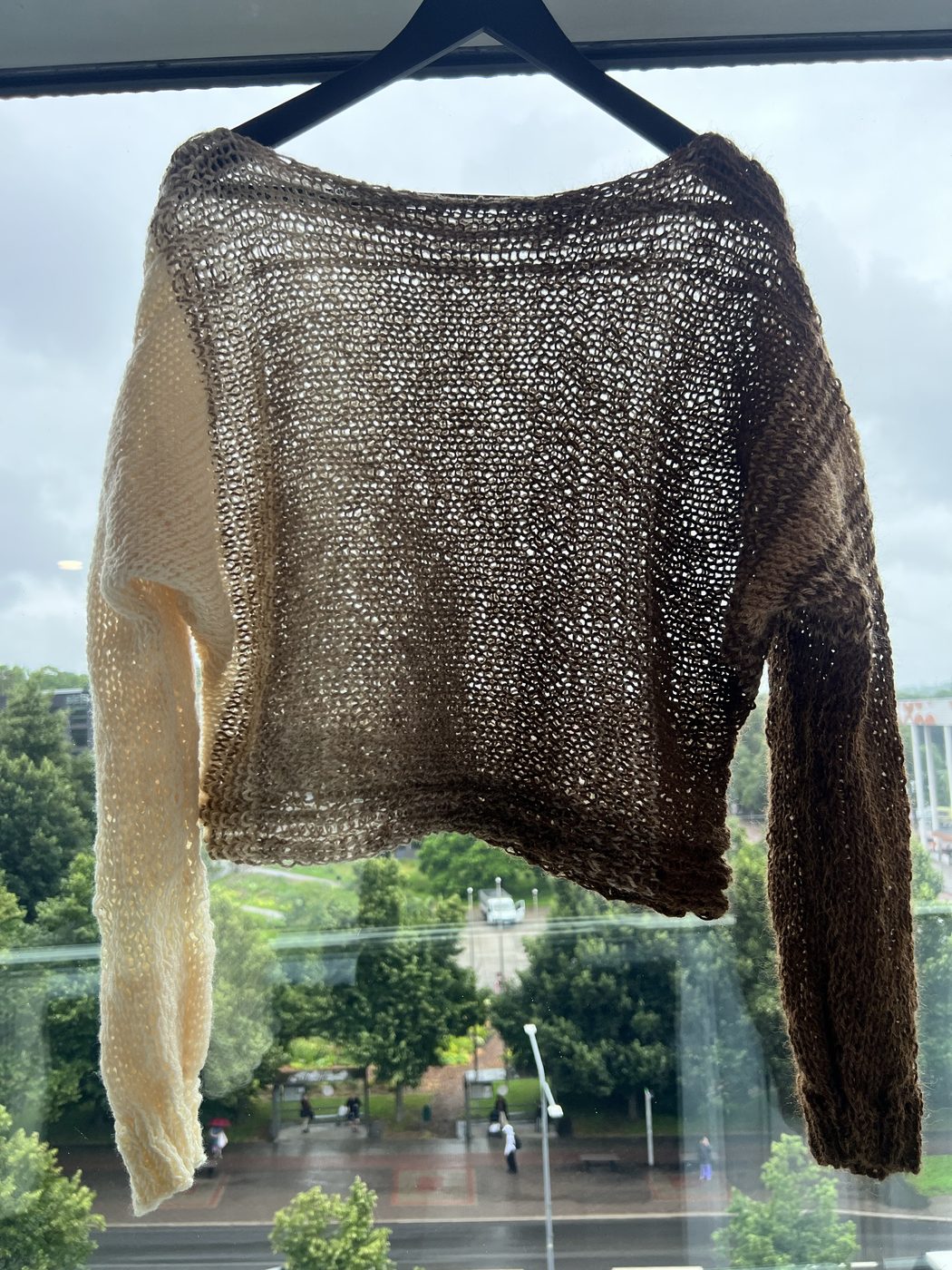 Beige Hali pullover on a hanger by a window, showing the airy loose-knit gradient from cream cuff through tan body to dark brown cuff