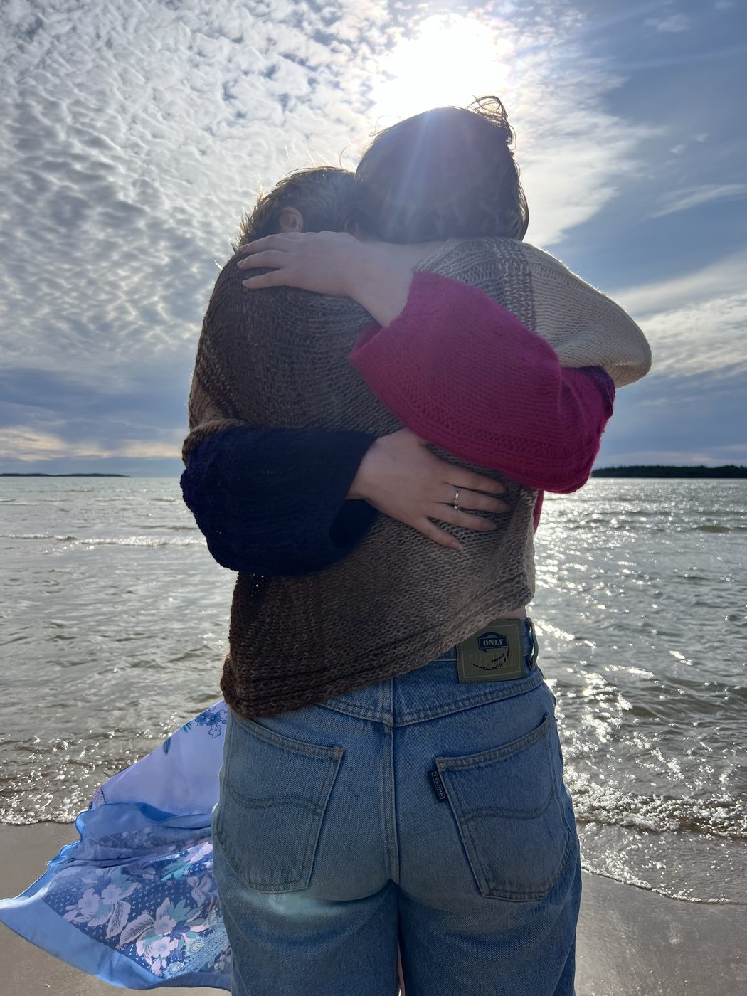 Two people hugging by the sea at sunset, both wearing Hali pullovers — one beige-to-brown ombre, one cream-to-pink. 'Hali' means hug in Finnish.