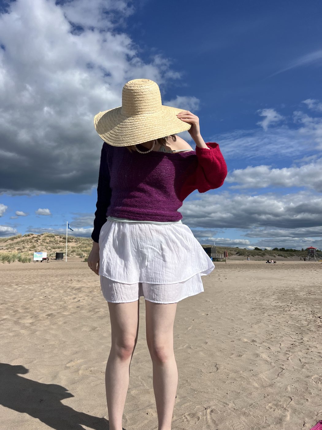 Model at Yyteri beach wearing a Hali pullover in a bold red-to-magenta-to-navy gradient, straw sun hat, white skirt, cloudy sky