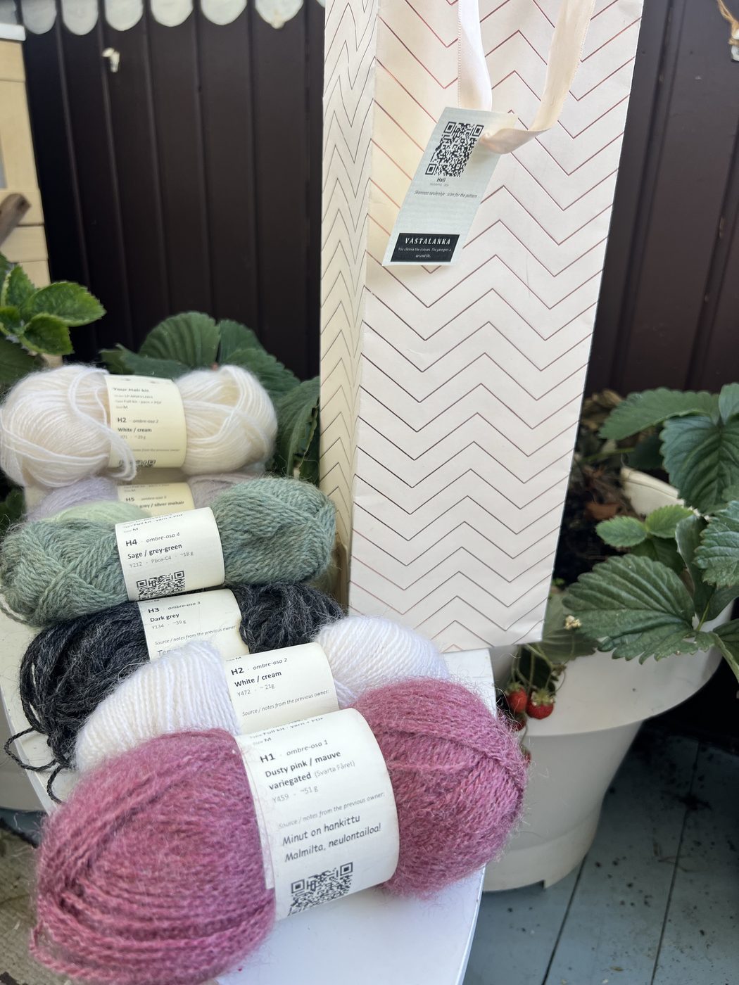 A Hali kit: six wound yarn balls in an ombre gradient from dusty pink through white, sage-green and dark grey, alongside a repurposed cream chevron gift bag with a Vastalanka QR hangtag