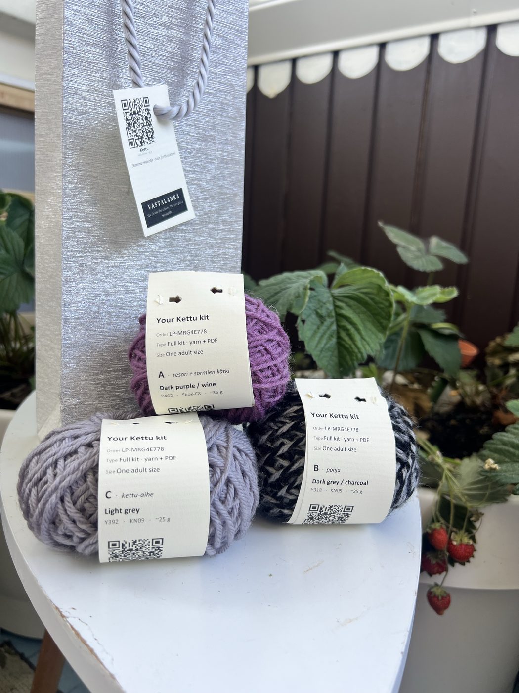 A Kettu kit: silver wine-bottle bag with a Vastalanka QR hangtag, and three wound yarn balls with printed ball-bands for slots A, B and C