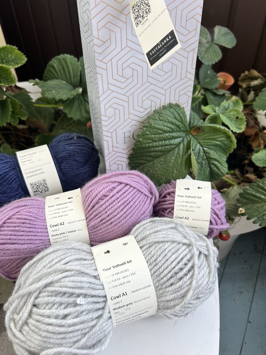 A Vohveli cowl kit: two wound yarn balls with printed bands for the body and waffle accent slots, in a repurposed paper bag with QR hangtag